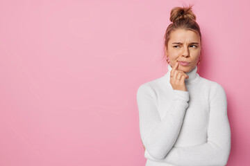 Horizontal shot of discontent sullen young woman looks thoughtfully away purses lips wears casual white turtleneck isolated over pink background with copy space for your promotional content.