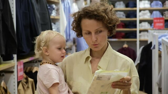 Young adult single mother with a capricious crying toddler on her clothing shopping in a fashion store, parenting routine
