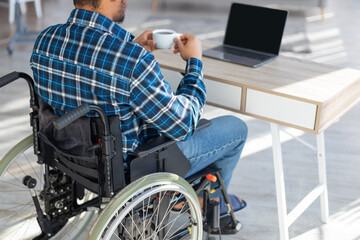 Unrecognizable handicapped black man in wheelchair using laptop for online work from home, having coffee, mockup