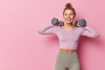 Athletic woman in sportswear raises dumbbells pumps biceps in gym being full of energy leads healthy lifestyle has happy expression isolated over pink background copy space for advertisement