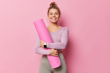 Fototapeta premium Pleased fitness trainers going to conduct yoga class embraces rolled karemat smiles gladfully has combed hair dressed in activewear uses smartwatch isolated over pink backgound. Time for training