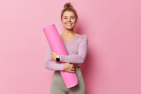 Pleased fitness trainers going to conduct yoga class embraces rolled karemat smiles gladfully has combed hair dressed in activewear uses smartwatch isolated over pink backgound. Time for training