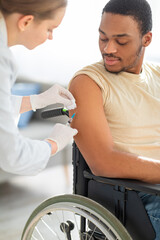 Young doctor giving injection of coronavirus vaccine to handicapped black man in wheelchair at home