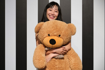 Young asian woman hugging a teddy bear standing on a striped room floor.