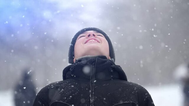 European teenage boy dressed in a black jacket outdoors in snowy weather in winter, slow motion. He looks up and rejoices