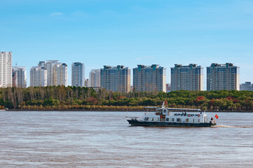 Chinese pleasure boat on the Amur River in autumn against the background of buildings in Heihe, China. View from the embankment of the city of Blagoveshchensk, Russia. - obrazy, fototapety, plakaty