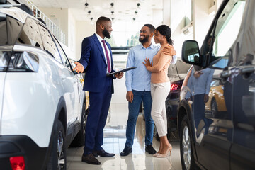Handsome Black Manager In Dealership Center Showing Car To Young Couple