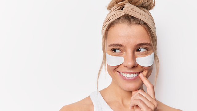 Care for your beauty. Positive dreamy young woman smiles happily applies anti fatigue mask under eyes moisturizes skin wears headband isolated over white background with blank space on left.