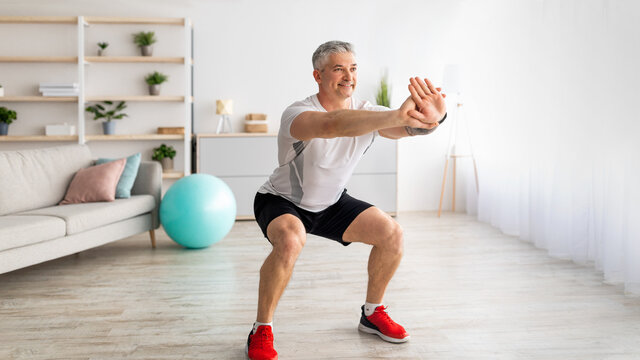 Strength workout concept. Active mature man doing squats at home, exercising in living room, panorama, free space