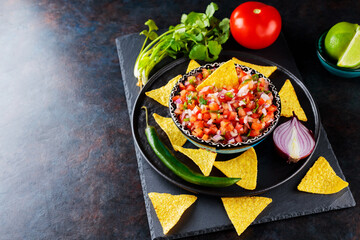Mexican Pico de Gallo salsa and ingredients. Bowl of tasty Pico de Gallo salsa and nachos on dark background. Top view. Copy space