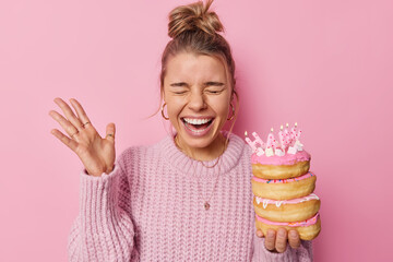 Overjoyed emotional woman exclaims gladfully keeps hand raise holds pile of delicious doughnuts celebrates birthday wears knitted sweater isolated over pink background. Time for celebration.