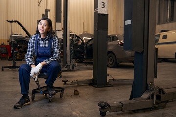 Fototapeta na wymiar Joyous lady in overalls resting on chair at repair shop