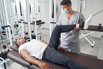Experienced physiotherapist applying gym equipment for patient leg rehabilitation - obrazy, fototapety, plakaty