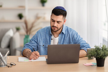 Focused israeli man working on laptop amd writing at home