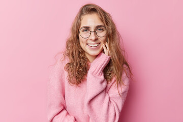 Portrait of lovely positive woman with long wavy hair smile toothily keeps hand on cheeks feels...