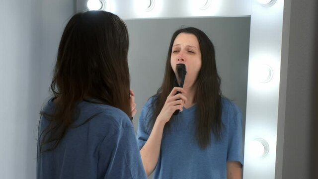 A young woman in front of a mirror is yawning and looks at her reflection. Tired sleepy woman with a hangover in the early morning. She tries to put her appearance in order.