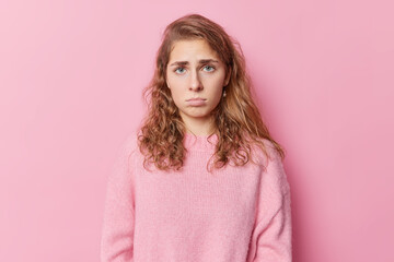Upset gloomy blue eyed woman has pity look feels discontent and offended looks dissatisfied pouts lips dressed in casual jumper isolated over pink background expresses sadness. Negative emotions