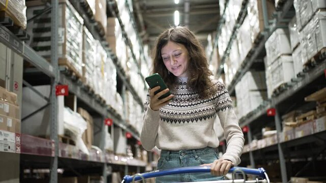 Brunette-haired young woman in jeans and sweater walks along boxes on metal racks with shopping trolley and scrolls social media using phone