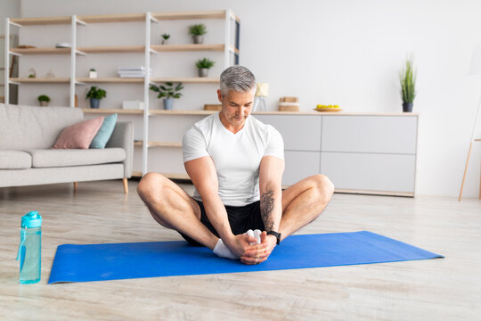 Fit mature man sitting in butterfly yoga pose on sports mat at home, doing flexibility exercises, copy space