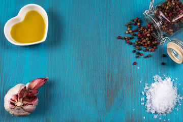 Blue wooden background with a small bowl of olive oil, next to a pot with peppercorns, a head of garlic and salt flakes. Copy space.