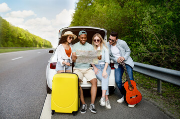 Multiracial young people sitting in car trunk, studying map, searching for their travel...