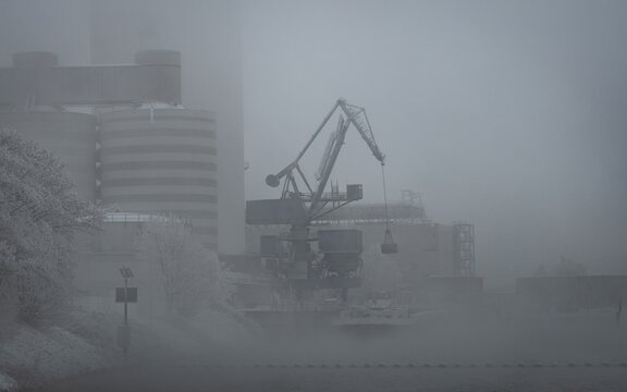 Bremen, Germany. First hoarfrost at Weserwehr weir next to swb Stadtwerke power plant beneath Weser river in foggy ice cold Hastedt. Chemical industry and moody bridges. luffing crane in front.
