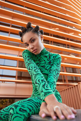 Vertical shot of serious beautiful woman dressed in green costume has bun hairstyle poses outdoors...