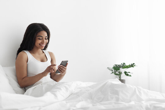 Happy african american woman blogger typing on smartphone or surfing in internet, sitting in bed, free space - Powered by Adobe