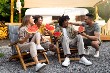 Joyful diverse friends chatting near camper van, eating yummy watermelon, sitting in lounge chairs,...