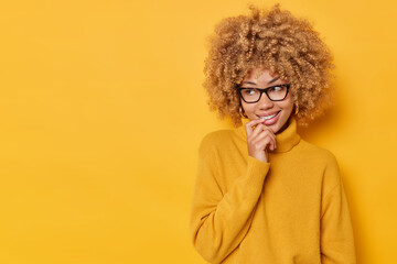 Fototapeta na wymiar Horizontal shot of dreamy lovely young woman keeps finger on lips looks away with curious happy expression dressed in comfortable jumper and spectacles isolated over yellow background copy space