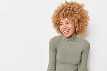 Joyful curly haired woman looks happily away laughs at something wears casual turtleneck isolated over white background copy space away for your promotional content. Positive emotions concept