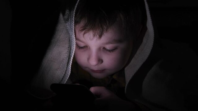 little cute boy sits at night with a phone under the blanket . Plays games.