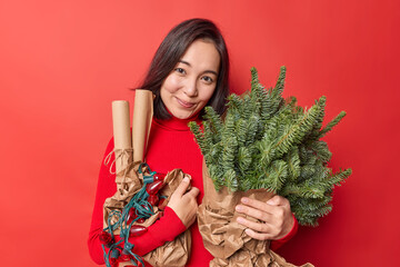 Pleased pretty Asian woman with dark hair carries holiday attributes for decoration awaits New Year dressed in casual turtleneck isolated over red wall. Lovely female model gets ready for Christmas