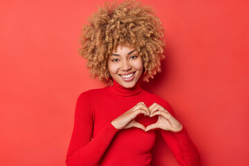Feminine young happy woman with curly hair cheerful smile shows heart sign looks tenderly at camera expresses love to someone stands in turtleneck against vivid red background. Be my valentine