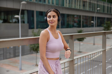 Outdoor shot of thoughtful healthy sportswoman listens music in headphones wears top and shorts checks time on smartwatch poses at bridge against city background concentrates away into distance