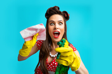 Crazy young pinup housewife wearing retro outfit and rubber gloves, holding spray detergent and rag over blue background