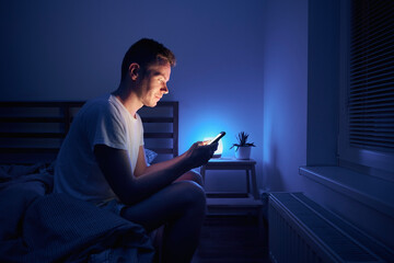 Lonely man sitting on bed in bedroom and using phone at night. Face illuminated from smartphone display.