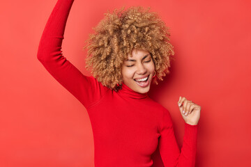 Delight of victory. Carefree curly haired young woman makes triumph dance shakes arms moves carelessly celebrates success enjoys achieving goalss wears turtleneck isolated over red background.