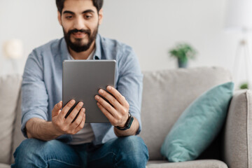 Millennial arab man sitting on sofa with tablet computer, working or studying online at home, focus on device