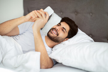Social networks, morning good news, positive message at weekend. Cheerful arab man waking up and looking at smartphone