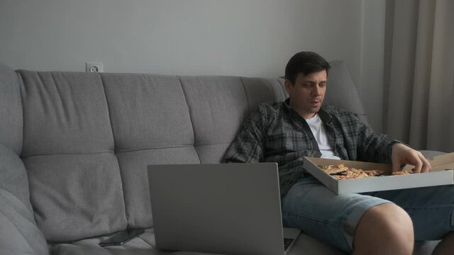 Young man is sitting on sofa eating slice of pizza from delivery cardboard box and watching video on laptop. Man is eating pizza with black dough. Modern european interior in apartment.