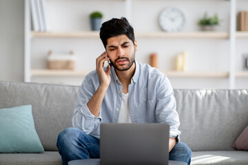 Remote business. Concerned arab freelancer talking on cellphone and using laptop computer, sitting on sofa at home