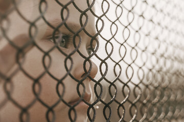 A little girl with a sad look behind a metal fence. Social problem of refugees and forced migrants