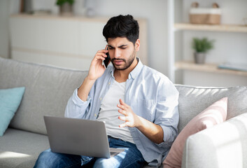 Computer problems. Displeased arab man talking on smartphone and looking on laptop screen at home