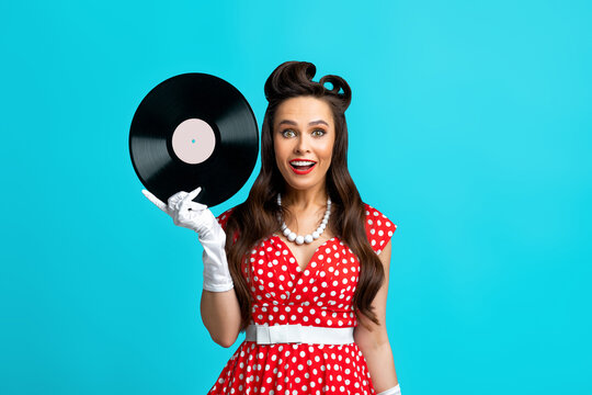Happy pinup woman in retro dress holding gramophone vinyl record, smiling and looking at camera over blue background
