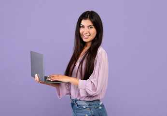 Portrait of confident armenian woman holding and using laptop computer isolated over purple studio...