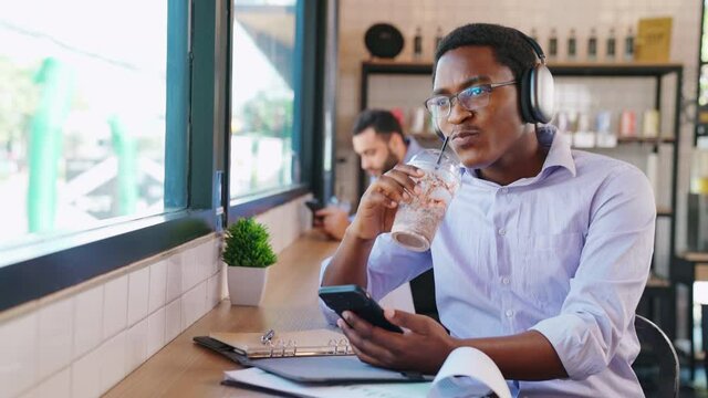 Attractive businessman listening music with headphones sitting near windows at coffee shop or cafe. Holding a plastic cup of ice coffee. Chill out life and digital lifestyle. Drinking and relaxation