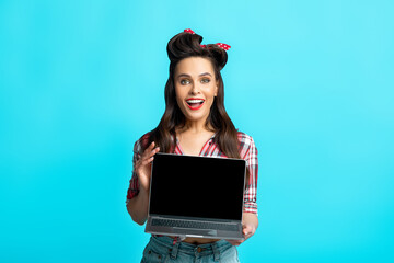 Smiling pinup woman in retro clothes showing laptop with empty screen on blue studio background,...