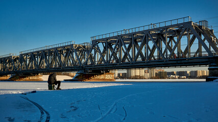 Fototapeta na wymiar A fisherman sits near the railway bridge over the Voronezh reservoir during the New Year's weekend. 