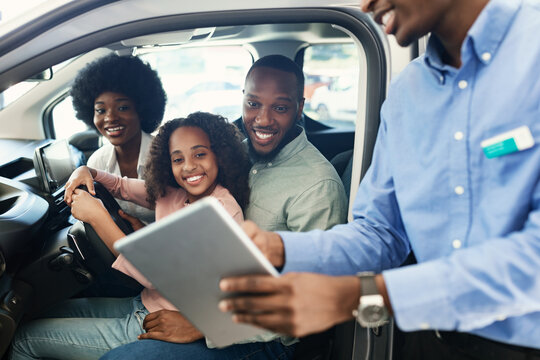 Black family testing new car, salesman showing them purchase or lease agreement on digital tablet at auto dealership